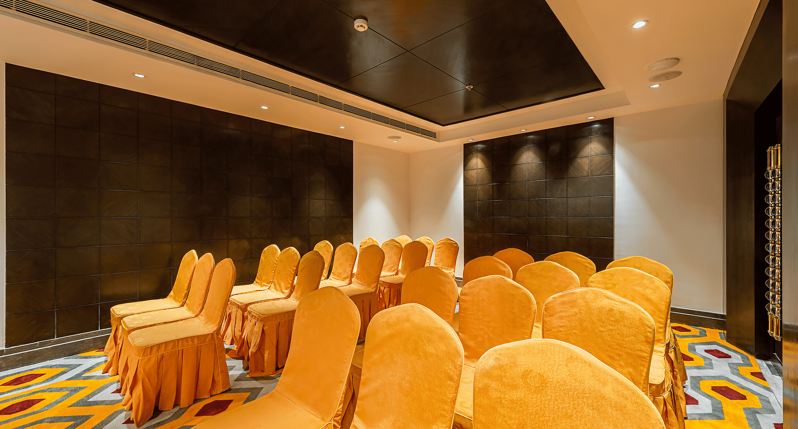 Conference room with yellow-orange chair covers, a dark tiled wall and a patterned carpet - TGI Grand Fortuna, Hosur