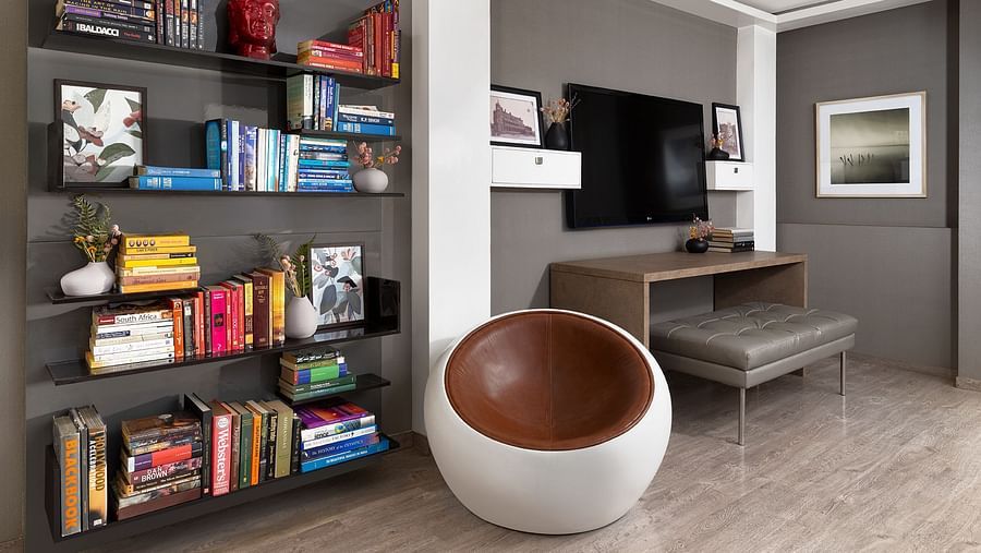 A contemporary living area at The Atara, Gurugram featuring a tall, dark bookshelf filled with books, a wall-mounted television above a sleek desk, and a distinctive circular armchair