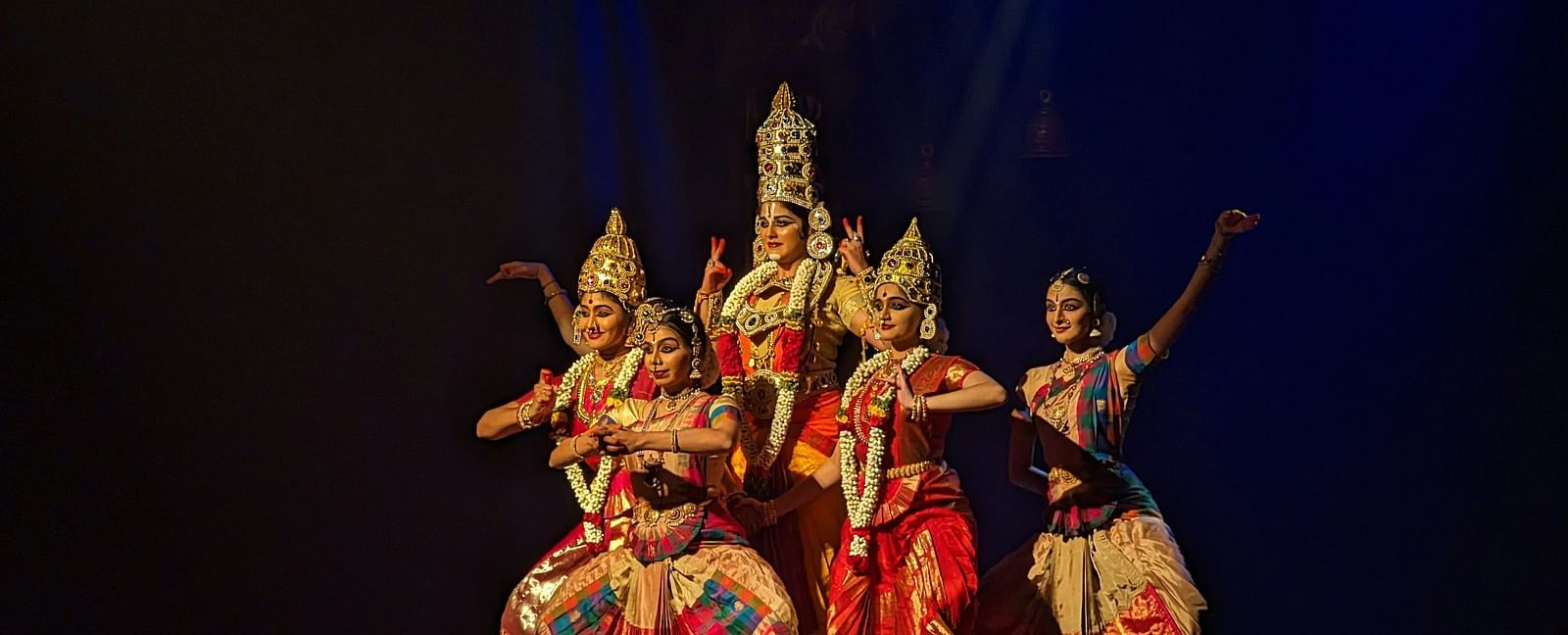 A group of women performing bharatanatyam on stage with a black background.