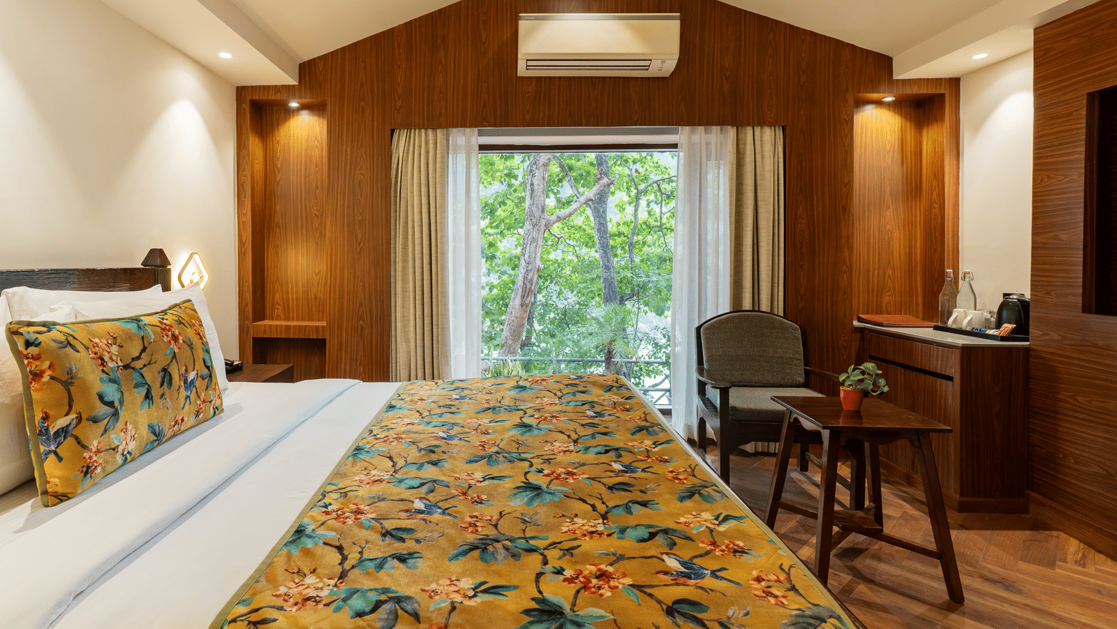 A bedroom featuring a bed, a television, a wooden chair and a table, a cabinet,  an A/C unit, and an attached balcony with a view of the trees outside | Summit by the Ganges Beach Resort & Spa, Rishikesh