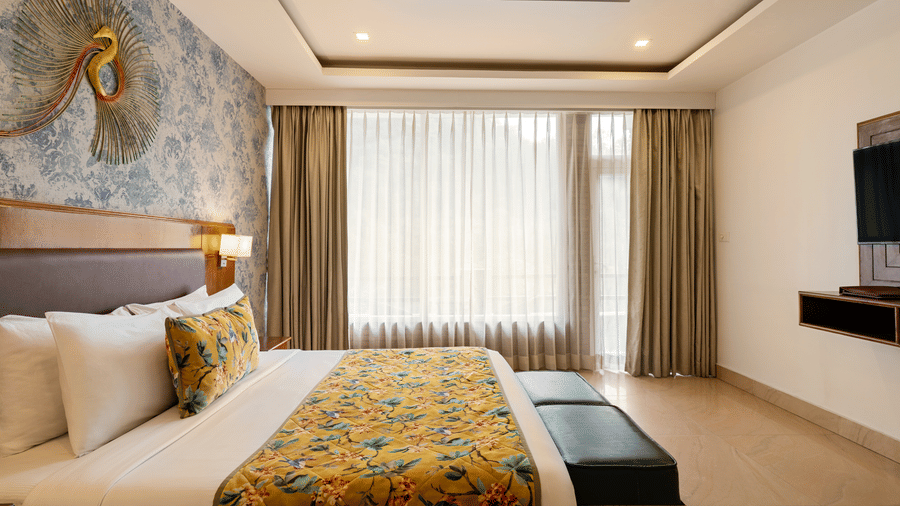A bedroom with a neatly made bed facing a television unit, a decorative art piece over blue printed wallpaper, and full-length curtains | Summit by the Ganges Beach Resort & Spa, Rishikesh