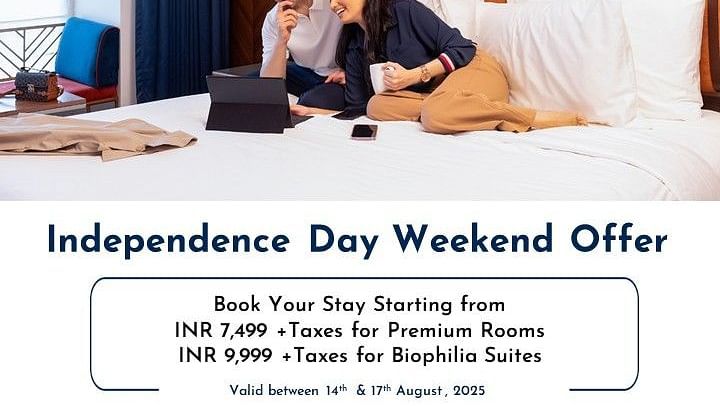 Creative hotel photography featuring a romantic couple relaxing together on a plush hotel bed while using a laptop, with Chennai's heritage architecture visible through the window, highlighting the cozy Independence Day weekend staycation experience.