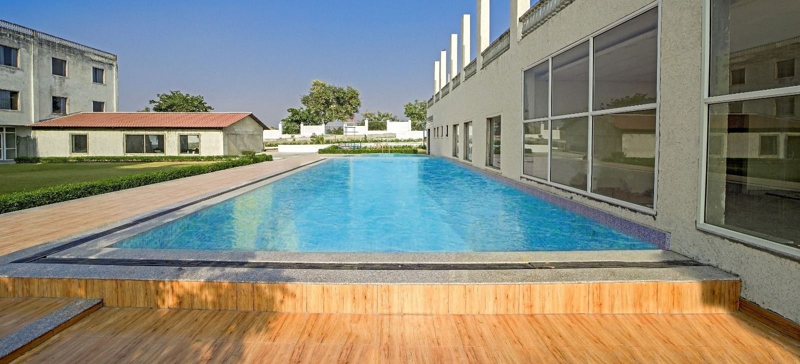Outdoor swimming pool at Grassfield Riveria Resort, Jaipur, surrounded by modern buildings and a wooden deck area.