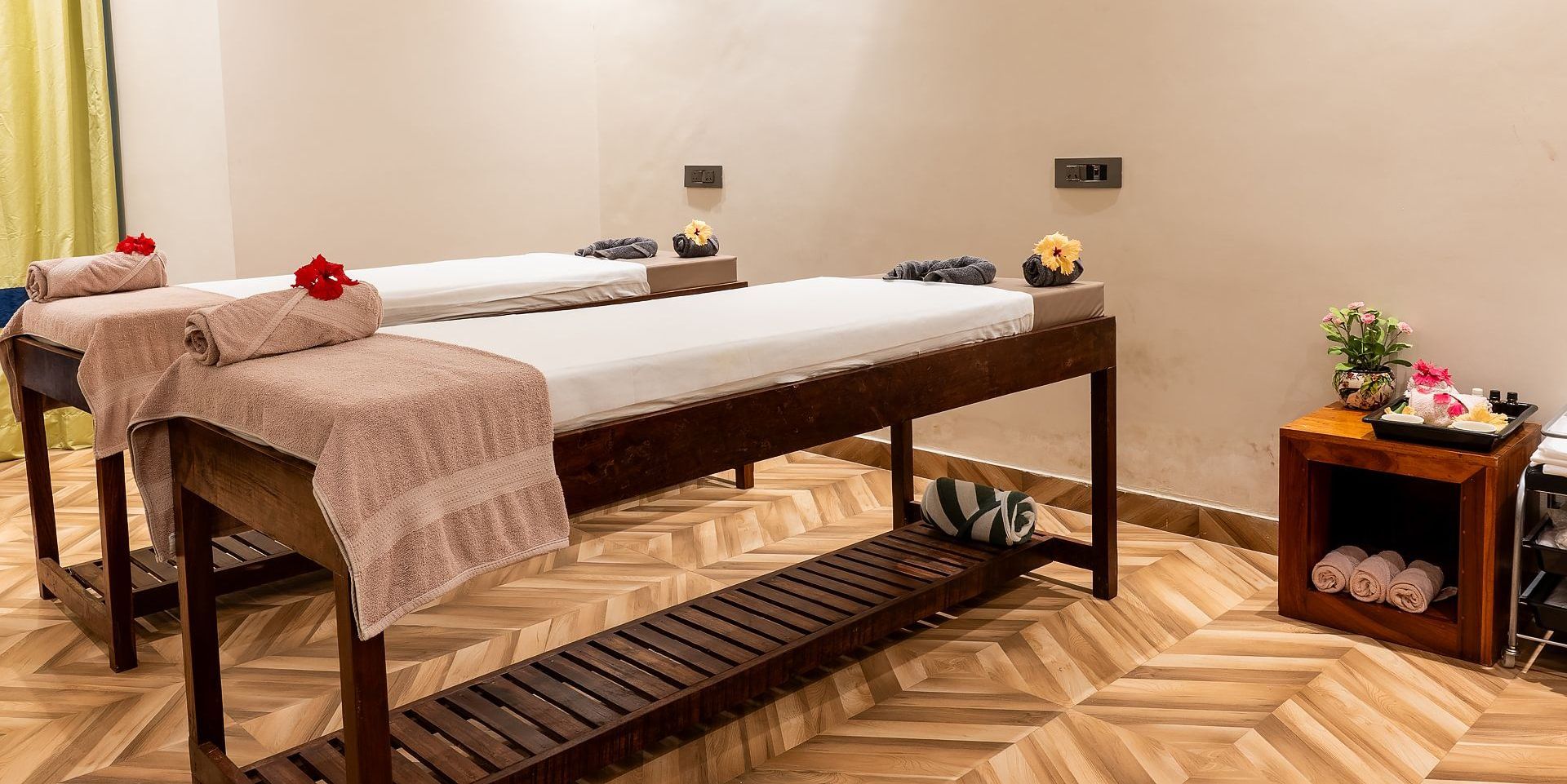 A bright couples spa room with two massage beds topped with white sheets, pink towels, and red flowers on a wood-patterned floor.