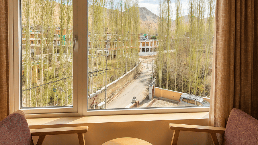 Close-up view through a large window showing mountains, greenery and a river below, framed by wooden armchairs and a round table set nearby.