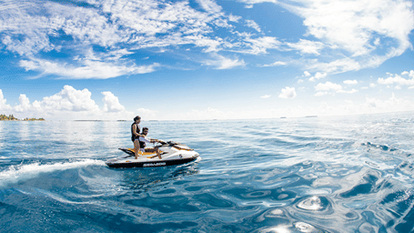A couple riding a white and orange jet ski across clear turquoise ocean waters under a sunny blue sky with scattered clouds.
