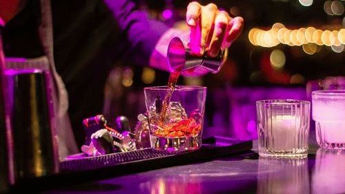 A bartender pouring a drink from a pouring cup into a glass with ice on a bar counter under ambient lighting.