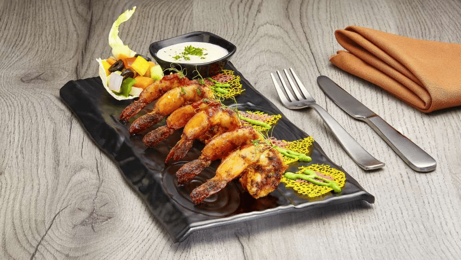Grilled prawn skewers plated on a black platter at The Citrine, Bangalore, with yogurt dip, vegetable garnish, fork, and knife on wood.