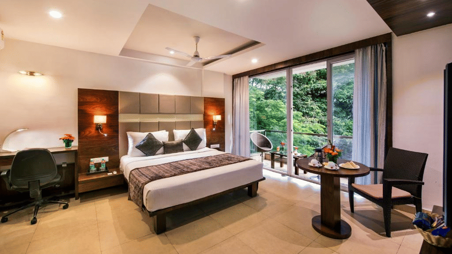 A hotel room with a king-size bed, work desk, and large windows overlooking greenery at Lords Eco Inn Jayanagar Bangalore.