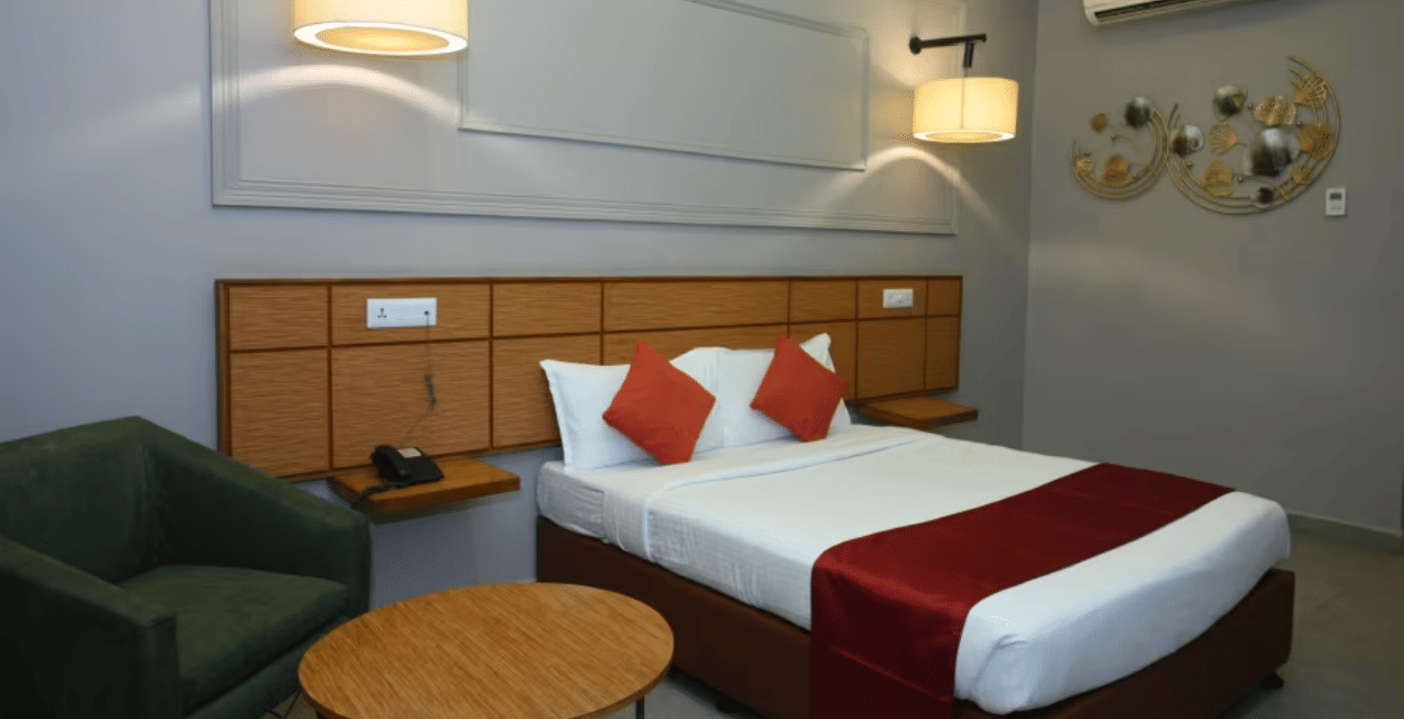 Bedroom with double bed, red runner, pillows, green chair, table and wall lamps at Max Hotel Prayagraj.