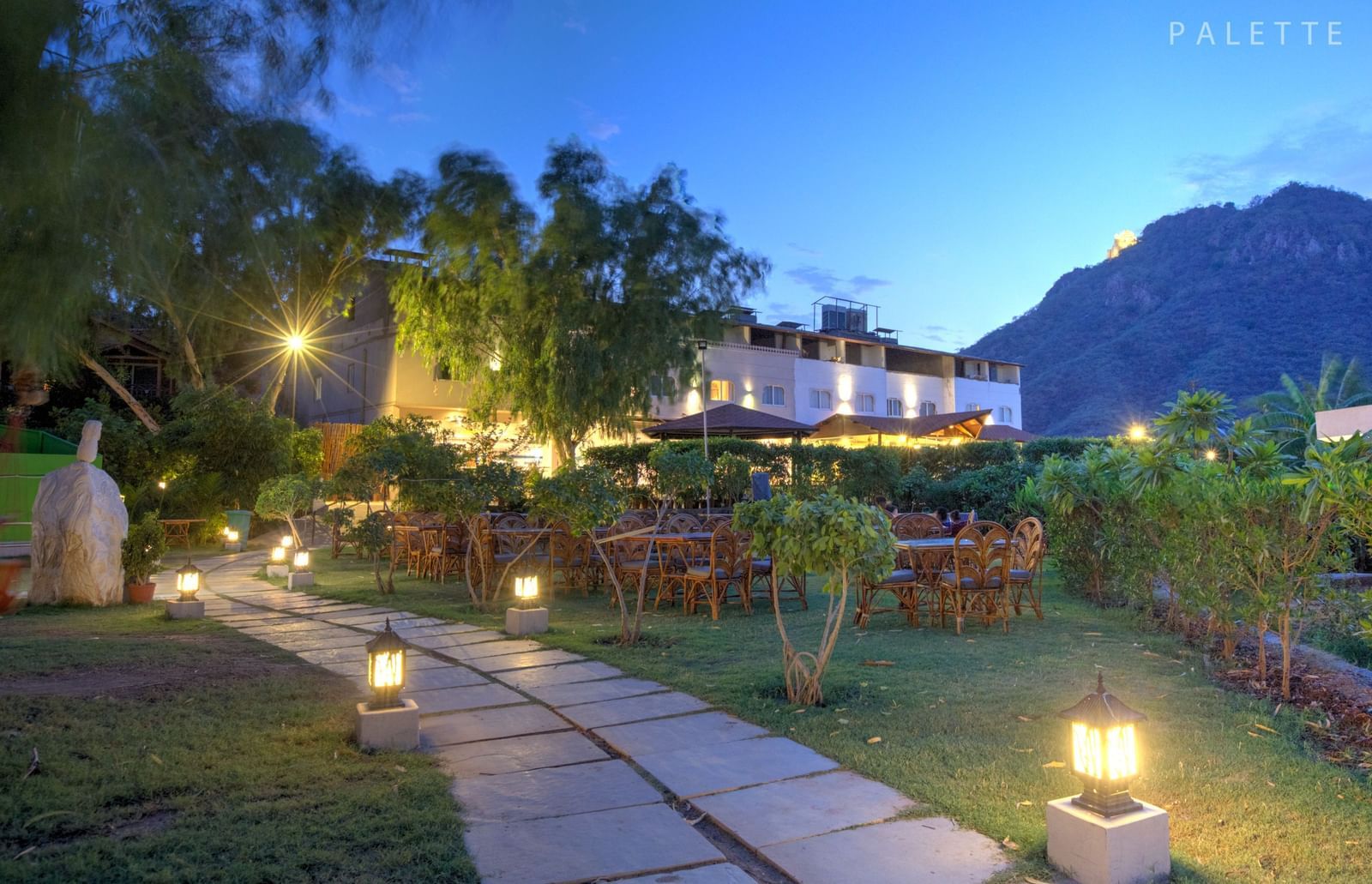 A pathway lit by lanterns leads through a garden at dusk, towards a building with warm lights.- Bamboo Saa Resort & Spa, Udaipur.