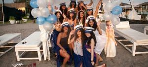 A group of women in blue and white attire, some wearing hats, pose outdoors under a decorated canopy with balloons and white lounge chairs.