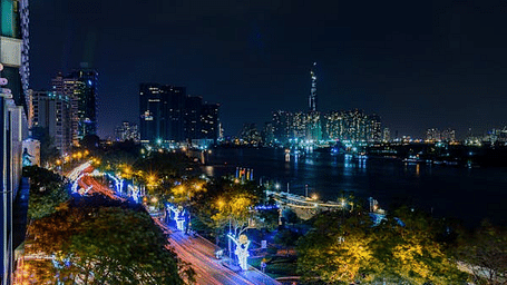 A nighttime cityscape view from a high vantage point, with a river, illuminated buildings, and trees visible.