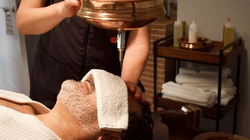  A man at YO1 Longevity & Health Resorts with a towel over his eyes is receiving a shirodhara treatment with warm oil poured onto his forehead