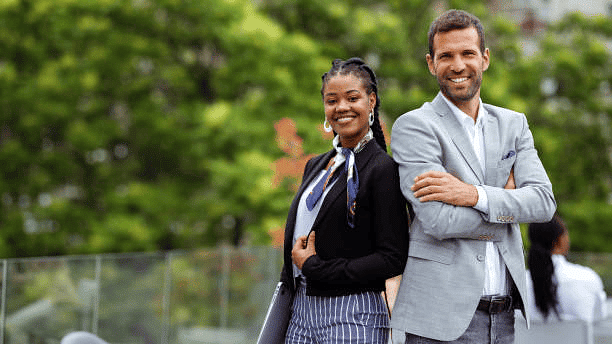 Stylish couple posing outdoors, dressed in formal wear with greenery in the background.