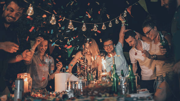 Friends enjoying a lively party with decorations, food, drinks, and joyful atmosphere under warm lighting.