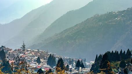A hazy, wide shot of a town nestled in a valley surrounded by large, misty mountains and evergreen trees in the foreground.
