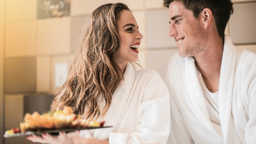 A smiling couple in robes enjoys breakfast in bed, sharing a joyful moment