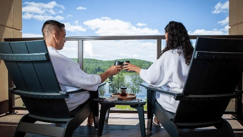 A couple in robes toasts with beverages while relaxing on a balcony with scenic views of green hills and a bright blue sky at YO1 Longevity & Health Resorts