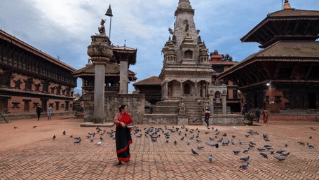 A woman in traditional attire walking among pigeons in front of ancient temples at Bhaktapur Durbar Square, Nepal.
