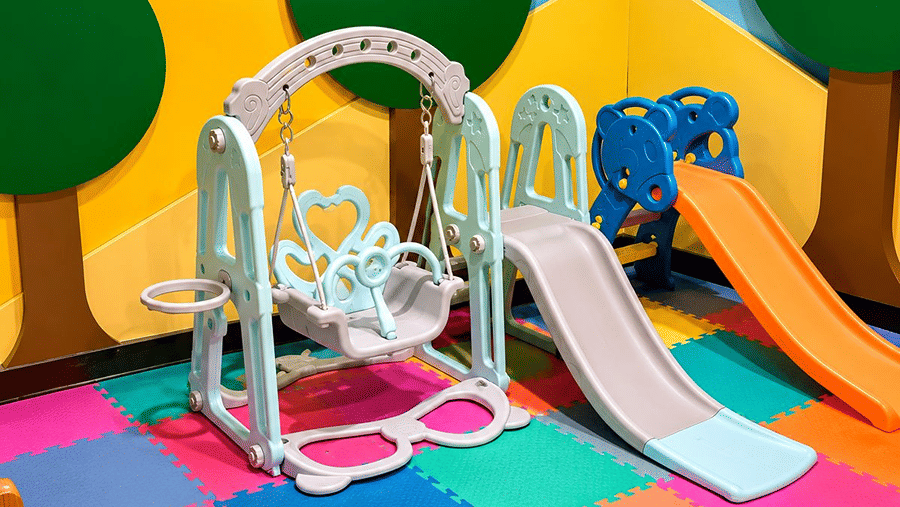 A kids play corner at Hotel Sonar Bangla, Mayapur, featuring a swing, a plastic slide, and a ring stacking toy.
