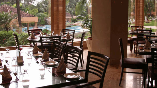 A restaurant dining area with wooden chairs and tables overlooking  a pool area framed by palm trees.