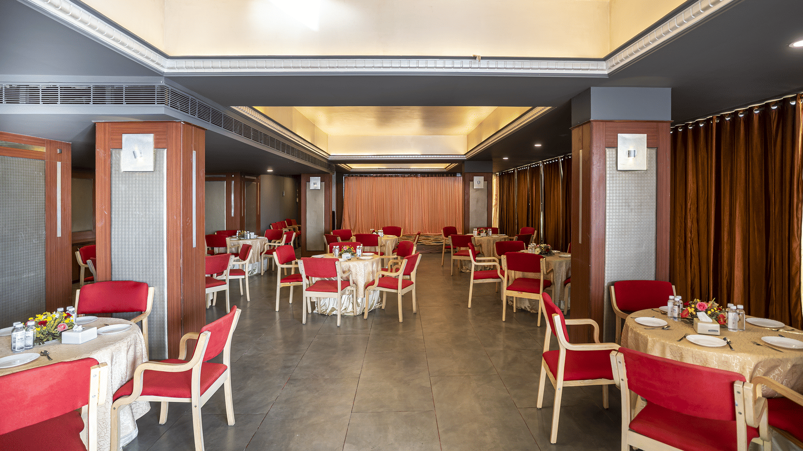 A dining hall with red chairs, white tables and warm ceiling lights at Infiniti Hotel, Indore.