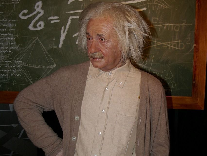 A wax figure of Albert Einstein standing in front of a black board with equations scribbled on it.