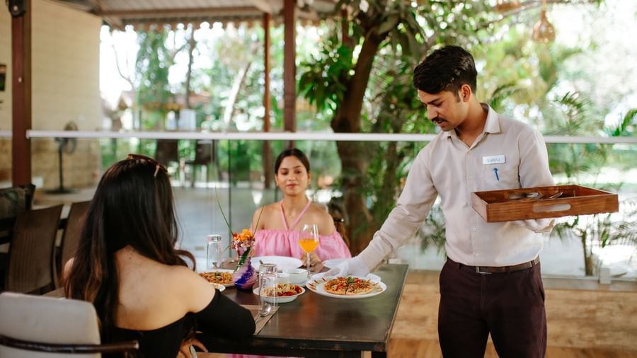 A waiter serving food with guests seated at a semi-open restaurant area with greenery in the background at Daksh Resort and Amusement Park.