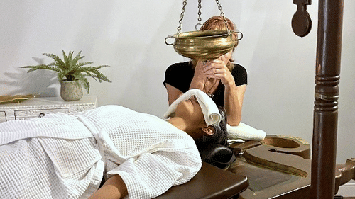 A person in a white robe lies on a wooden table, receiving an Ayurvedic shirodhara treatment with oil dripping onto their forehead at YO1 Longevity & Health Resorts, Catskills
