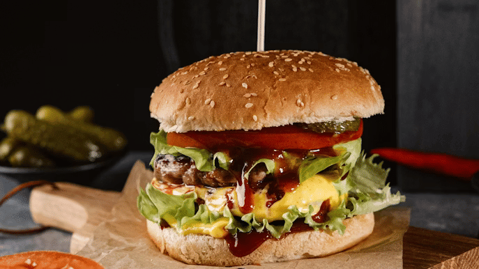 Cheeseburger on a sesame bun with lettuce, tomato, melted cheese, and sauce, served on a wooden board with pickles and tomato slices nearby.