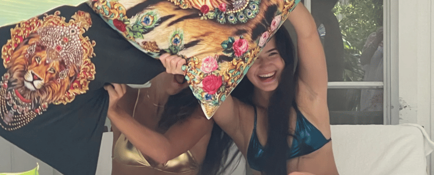 Two women in metallic bikinis are seated outdoors on white lounge chairs, playfully engaging in a pillow fight with large, decorative pillows featuring a tiger and lion print, enjoying their time at Casa Morada.