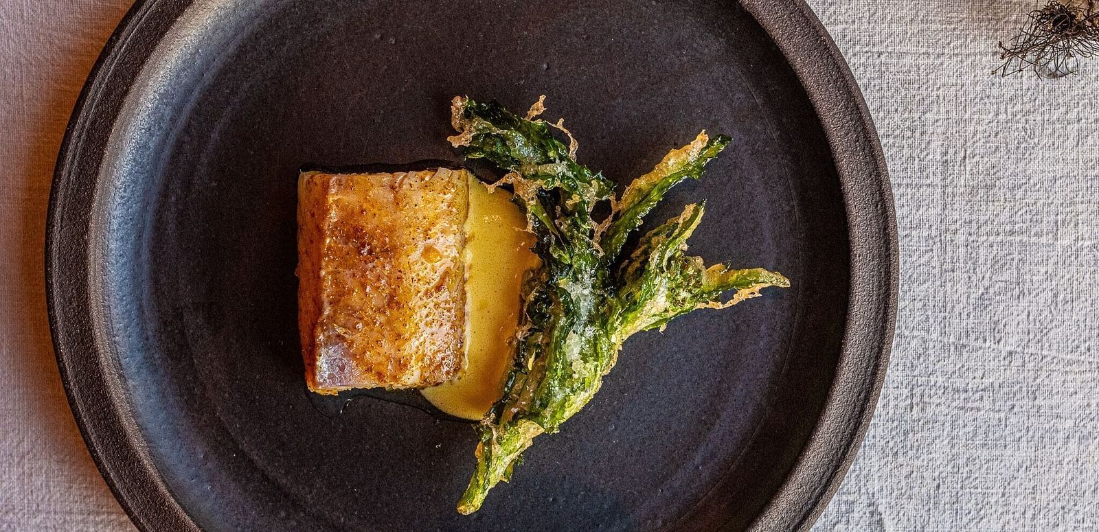 A dark plate holds a food item with textured sides and a side of leafy greens. 