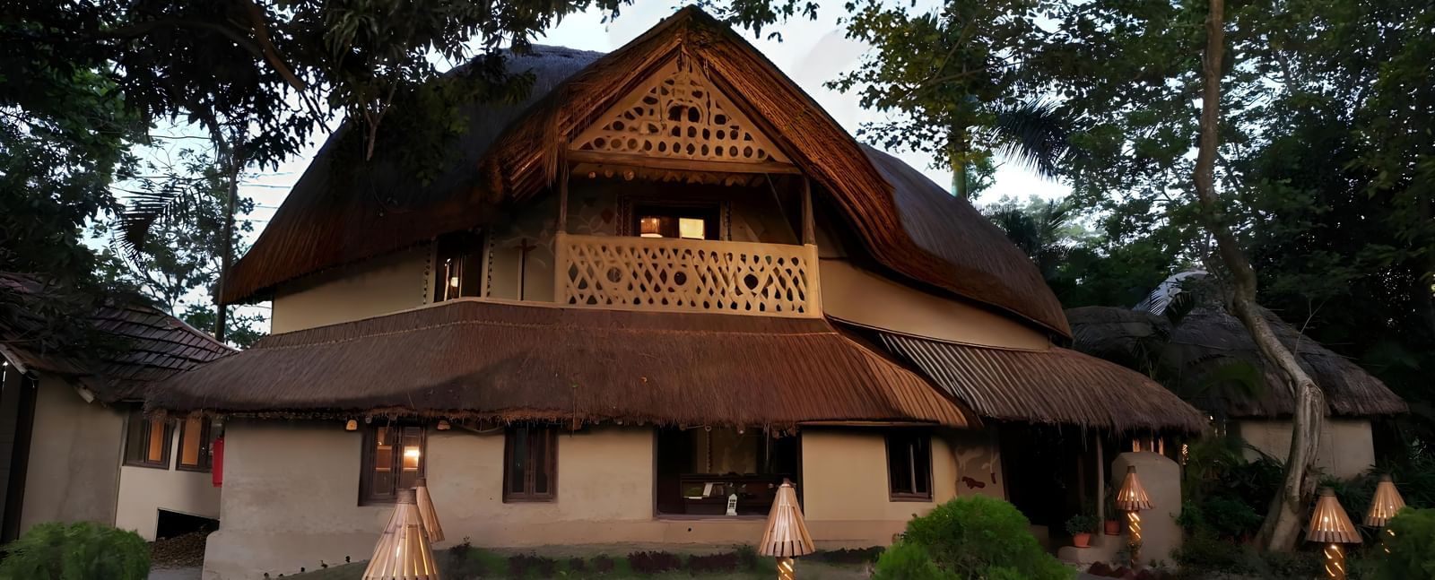A traditional wooden house with a tiled roof is situated amidst lush greenery | Vedic Village Spa Resort, Kolkata