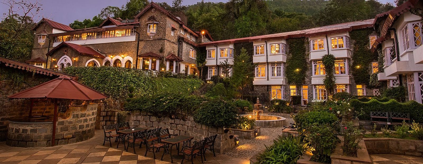 A scenic view of a the naini retreat, nainital, The resort features multiple buildings with stone exteriors and white trim. The surrounding landscape includes mountains, trees, and a winding pathway.