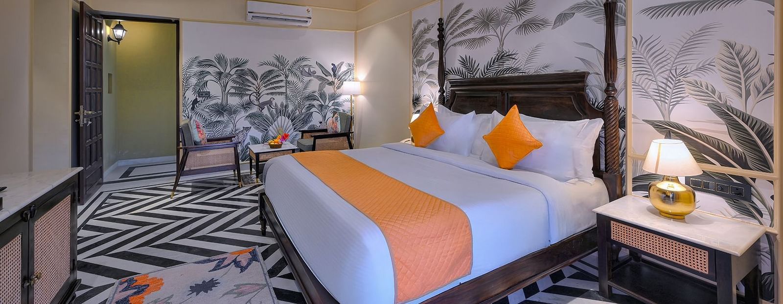 Superior Cottage featuring a four-poster bed with orange accents, chevron-patterned flooring, tropical wallpaper, a seating area, and a nightstand with a gold lamp - Singhvilas, Ranthambore