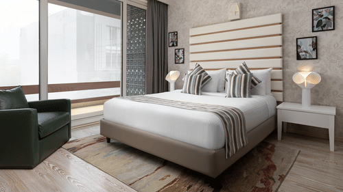 A stylish room at The Atara, Gurugram boasts a comfortable bed with a patterned runner, a plush green armchair, and a large window that offers a scenic outdoor view