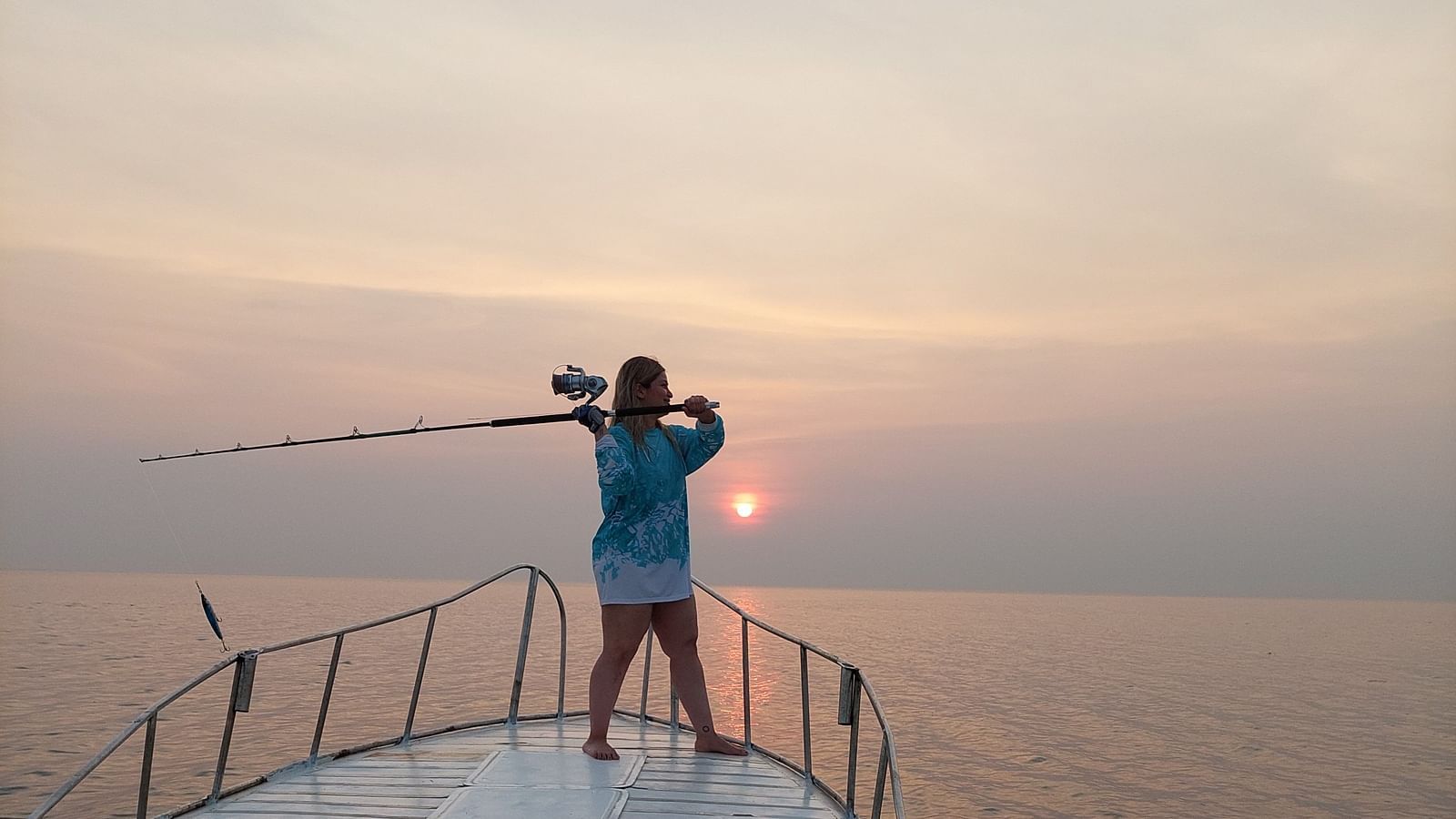 A lady holding a fishing rod over her shoulders standing on a yacht, with sun setting in the background.