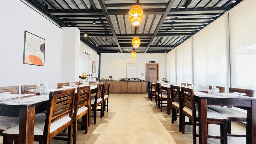Spacious restaurant dining area at Hotel Kyzen Smart Stays Madhapur with long wooden tables, chairs, pendant lighting, dark ceiling beams, and bright natural light.