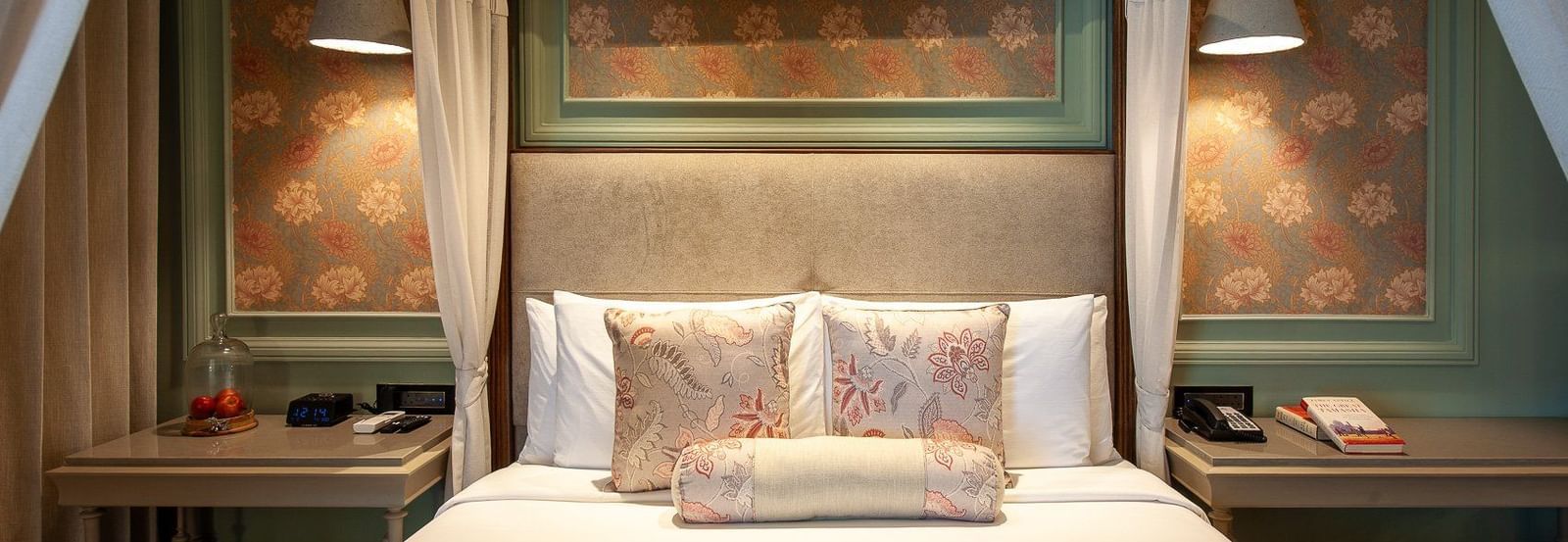 A close-up, front-facing view of the beautifully appointed four-poster bed in a suite at The Evren, Vagator. The bed is adorned with decorative pillows, and the background features elegant wallpaper and unique hanging lights.