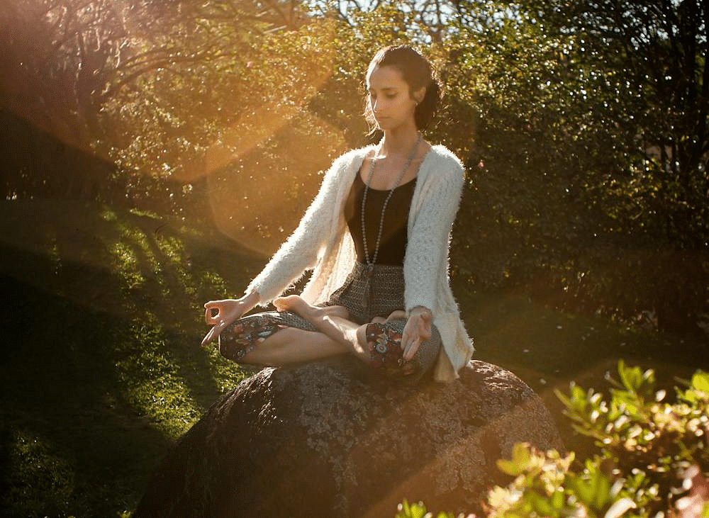 A woman sitting on a rock meditating in a lush green garden with bright sun rays falling on her.