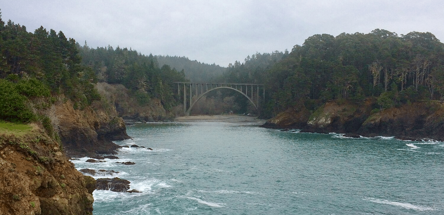 A concrete bridge arches over a calm river surrounded by rocky cliffs and dense evergreen forests under a cloudy sky.