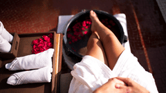 An image of a woman's leg resting in a round tub filled with water and rose petals, with towels neatly arranged on a tray beside it.