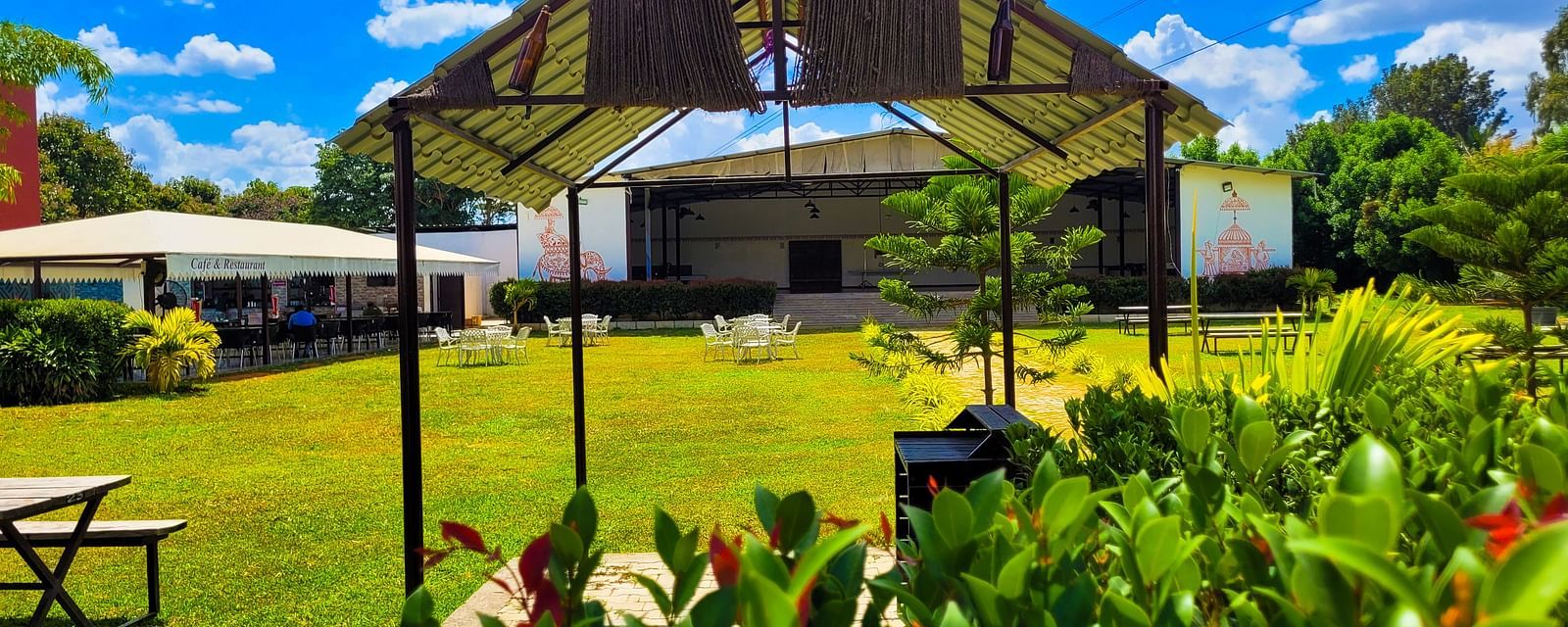 Covered area with tables lined under a beige canopy amidst lush greenery. | Vistar Resort, Bengaluru