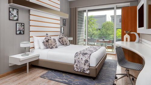 A modern bedroom at The Atara, Gurugram featuring a neatly made bed with a floral runner, a stylish white desk with an armchair, and a large window offering a refreshing view of lush greenery