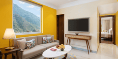 A cosy living room with a beige sofa, patterned pillows, a white oval coffee table, a wooden side table with a lamp, a wall-mounted television, and a large window offering a mountain view, with an open doorway leading to a bedroom - Aloha On the Ganges.