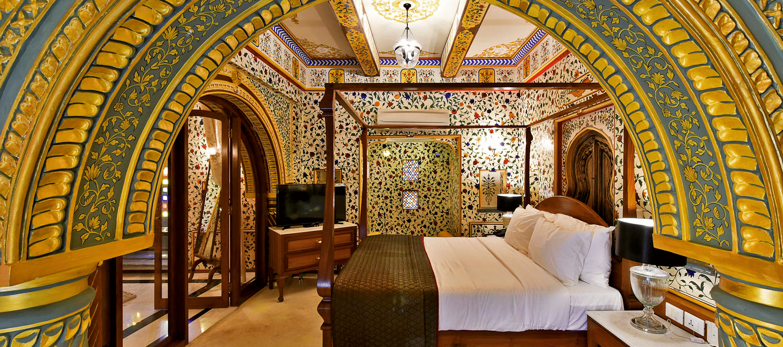 A luxurious palace bedroom at Gogunda Palace framed by an ornate gold and blue arch, featuring a wooden 4-poster bed and opulent decor.