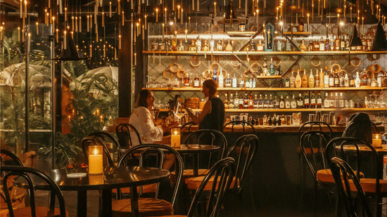 A warmly lit bar interior at The Spirit Bar featuring a backlit liquor shelf guests seated at the counter hanging pendant lights dark furniture and lush greenery in a moody atmospheric setting.