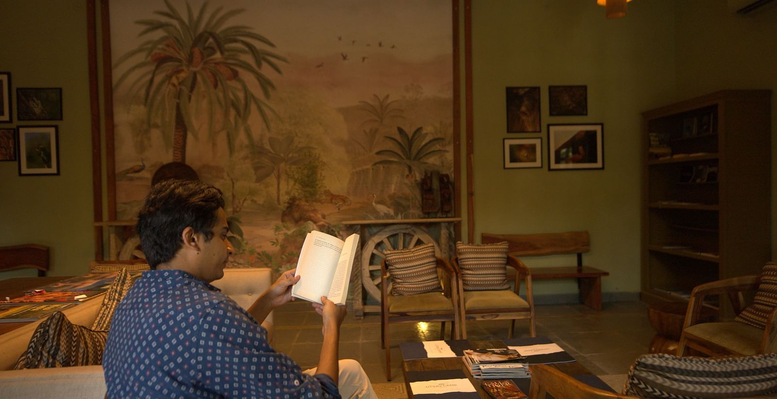 An image of a man reading a book sitting in a room which is well decorated and brightly lit