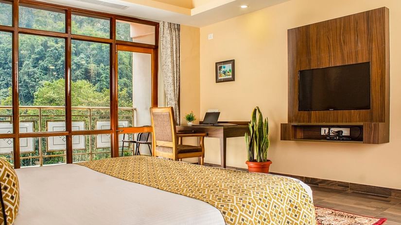 A side view of a bed in a room with glass window, writing desk and a TV on the wall | Sumi Yash Shree Hotels & Resorts